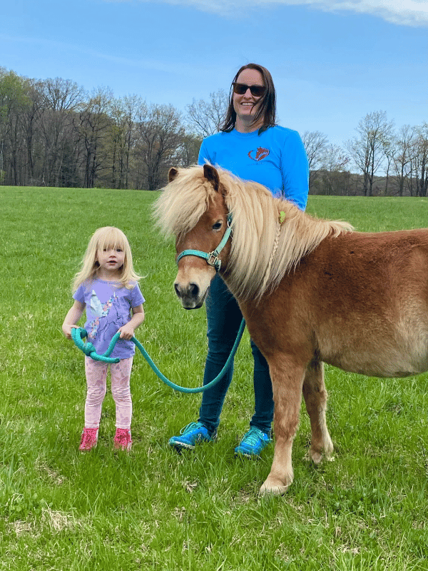 Little girl holds the rope of a chestnut pony, she is looking up at the pony. Amy Hand, Equine Gestaltist is holding the pony with her.